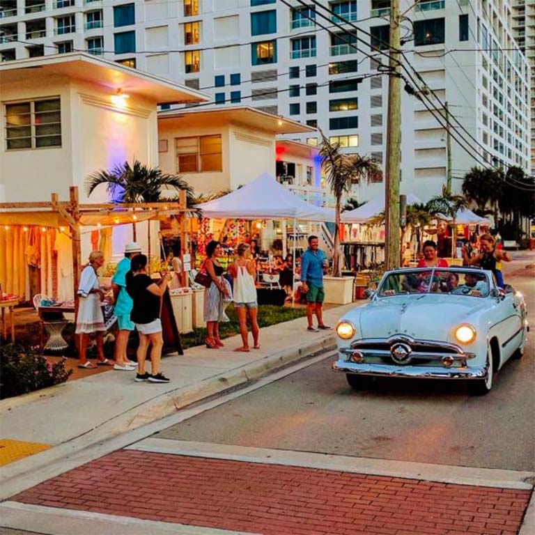 a white car drives down a street with people standing on the sidewalk in front of a building