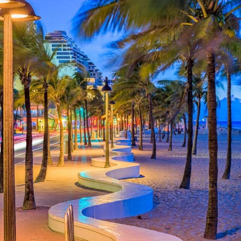 A beachfront promenade with palm trees, benches, and streetlights at dusk.